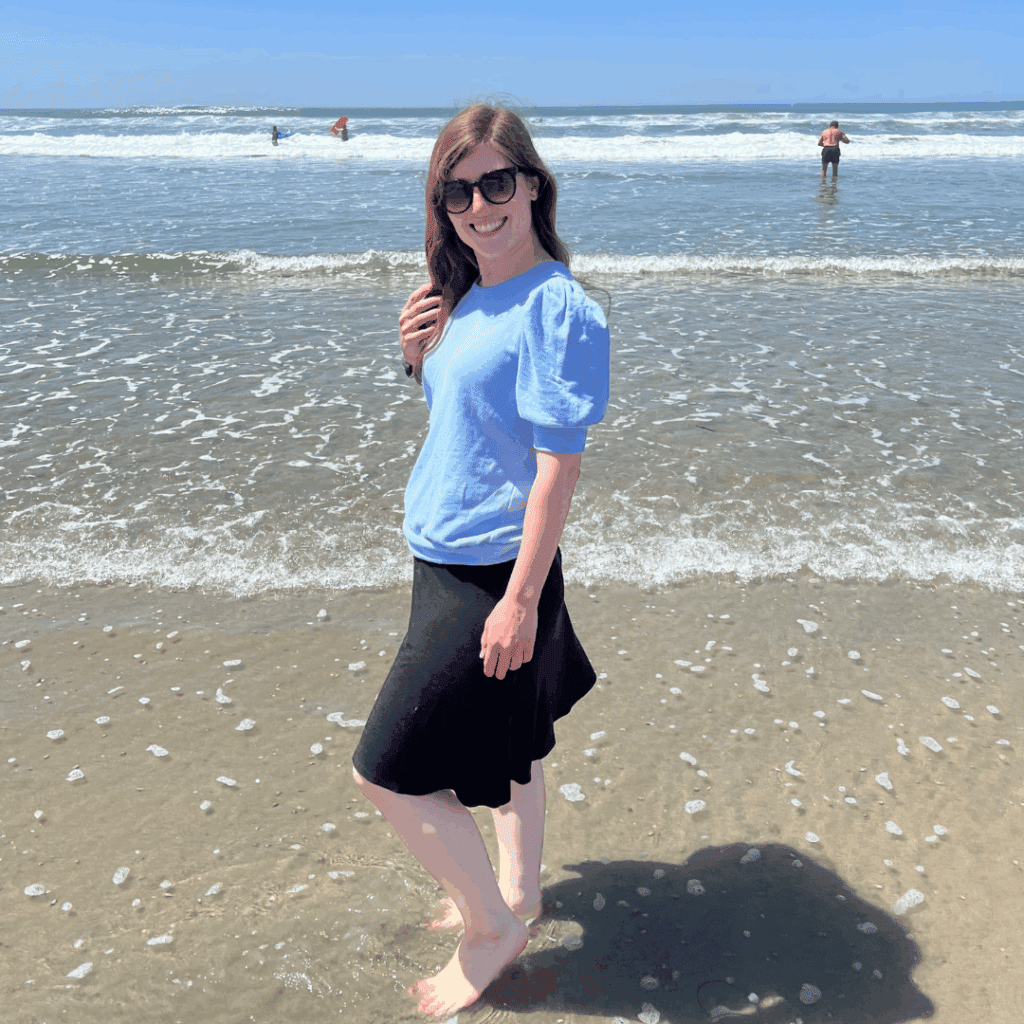 A woman in sunglasses, a light blue shirt, and a black skirt stands barefoot on a sandy beach with waves in the background, under a clear blue sky.
