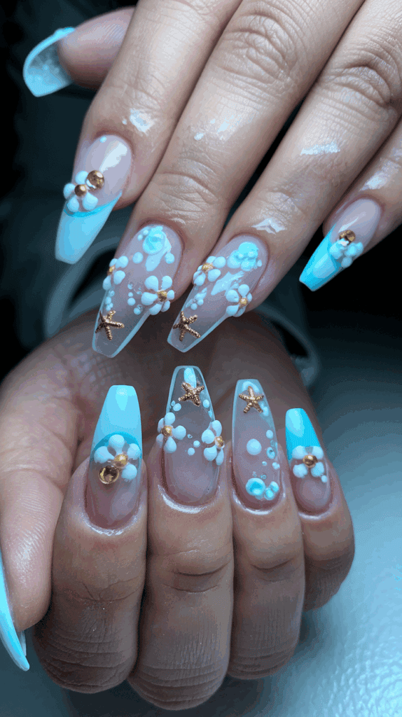 A close-up of a hand with long, almond-shaped nails featuring an intricate design. The nails have a glossy finish in a transparent base, decorated with white daisies and gold starfish embellishments. There are also blue and gold accents resembling bubbles and gold beads toward the tips of the nails, which are painted in a light blue color.