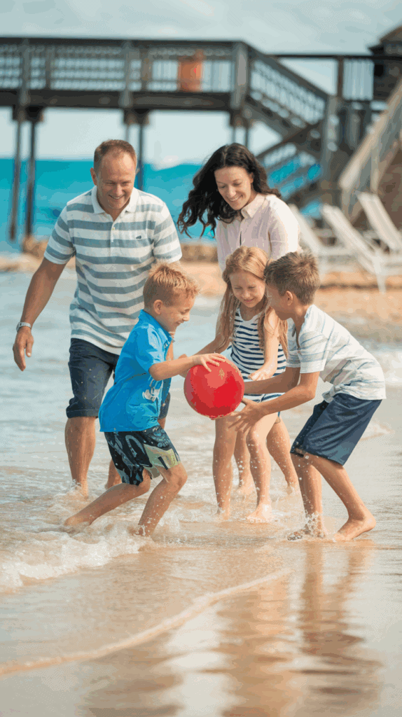 A family plays with a red ball on a beach, with three children in swimwear splashing in the shallow water and two adults laughing nearby. A wooden pier extends into the ocean in the background.