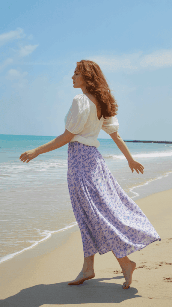 A woman with red hair walks barefoot along a sandy beach, wearing a white blouse and a purple floral skirt, with the ocean and blue sky in the background.