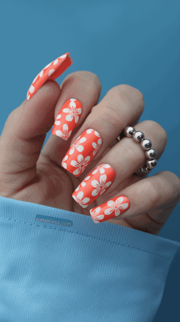 A hand with long, bright orange nails featuring white floral designs, resting on a light blue surface. The person wears a silver beaded ring on their middle finger.
