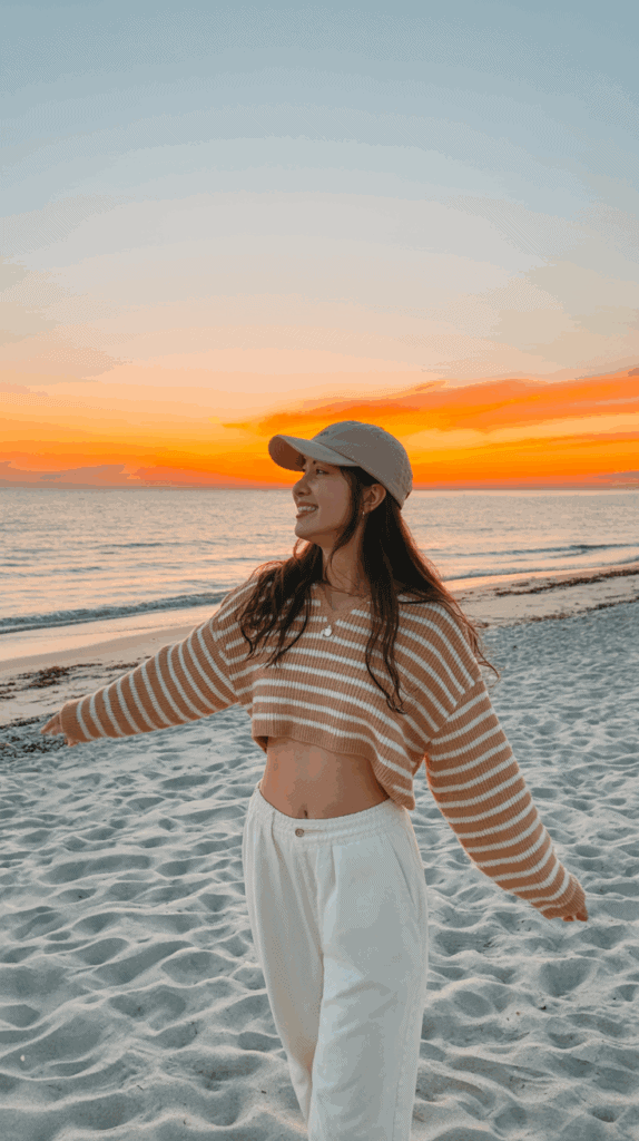 Person wearing a striped crop sweater and a cap, standing on a beach during sunset with arms outstretched.
