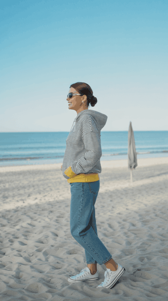 A woman wearing sunglasses, a striped hoodie with yellow accents, and jeans stands on a sandy beach with the ocean and a closed beach umbrella in the background.