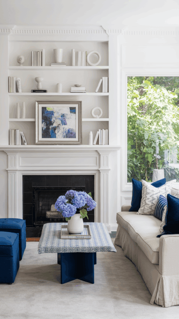 A stylish living room with a white fireplace and built-in shelves containing books and decorative items. Blue and white artwork is displayed above the fireplace. A beige sofa with blue and patterned pillows sits next to a blue ottoman. A coffee table with a blue patterned top holds a vase of blue hydrangeas. A large window offers a view of green foliage outside.