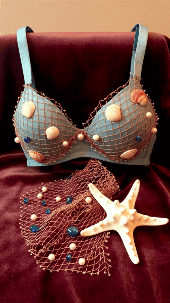 A decorated bra featuring blue cups overlaid with a net adorned with seashells, pearls, and blue beads, set against a dark fabric background. A matching netted accessory and a starfish are placed in front of it.