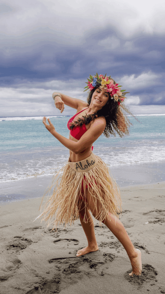 A woman in a red outfit and a grass skirt with "Aloha" written on it dances on a sandy beach. She wears a colorful flower crown and bracelet, with the ocean and cloudy sky in the background.