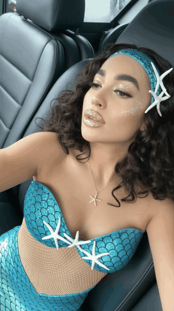 Person dressed in a mermaid-inspired outfit with a blue scale-patterned top adorned with starfish and glittery makeup, sitting in a car.