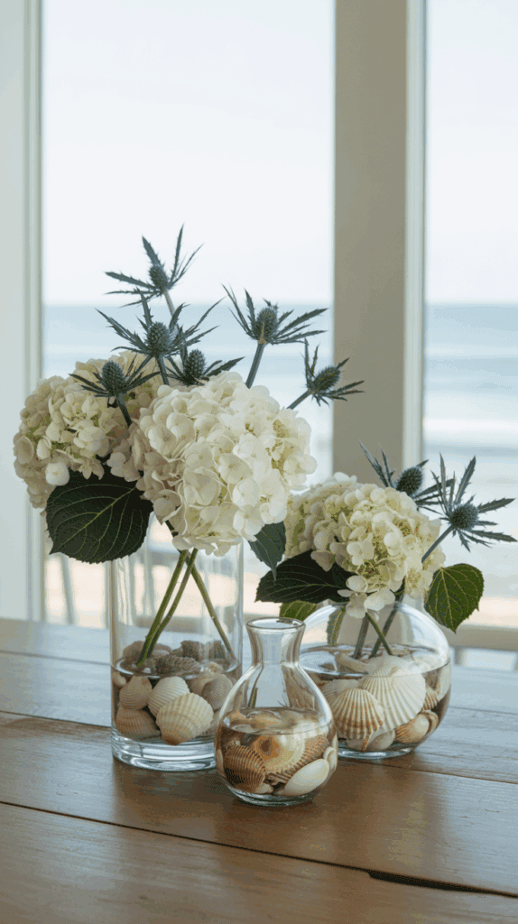 Glass vases filled with white hydrangeas and sea holly flowers, arranged with seashells at the bottom, set on a wooden table with a beach view in the background.