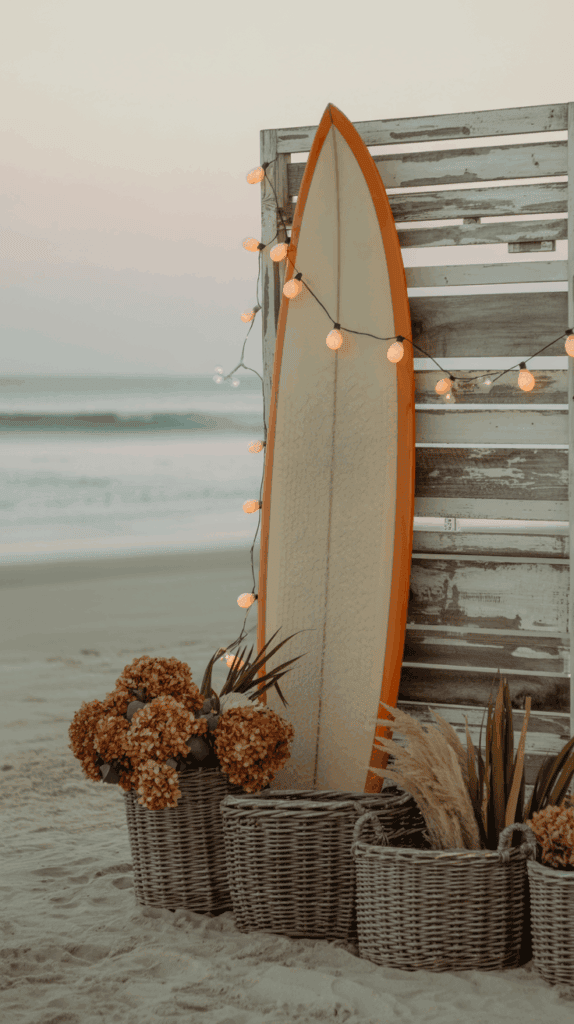 A surfboard with an orange edge leaning against a wooden fence adorned with string lights, surrounded by wicker baskets filled with dried flowers and grass, set on a sandy beach with the ocean in the background.