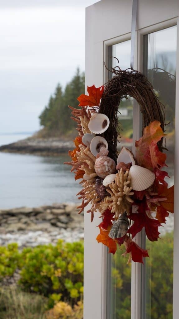 A decorative wreath made of seashells, coral, and autumn leaves hangs on a glass door, with a scenic view of the ocean and forested coastline in the background.