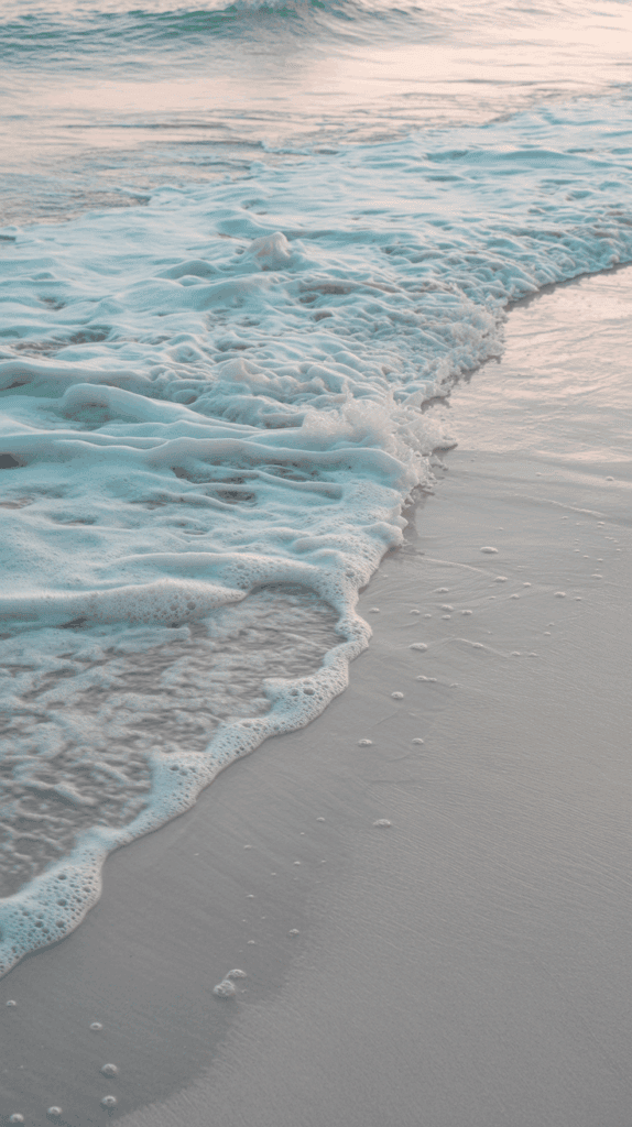 Gentle waves with foamy edges washing over a sandy beach at sunset, with soft pink and blue hues reflecting on the water and sand.
