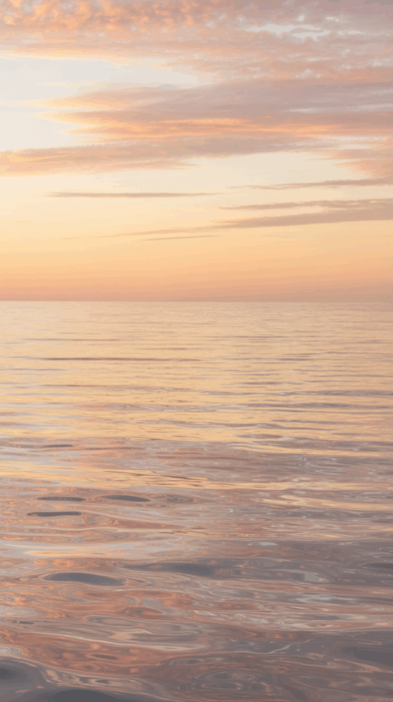 A serene view of the ocean at sunset with gentle orange and pink tones reflecting in the calm water, and soft clouds scattered across the sky.