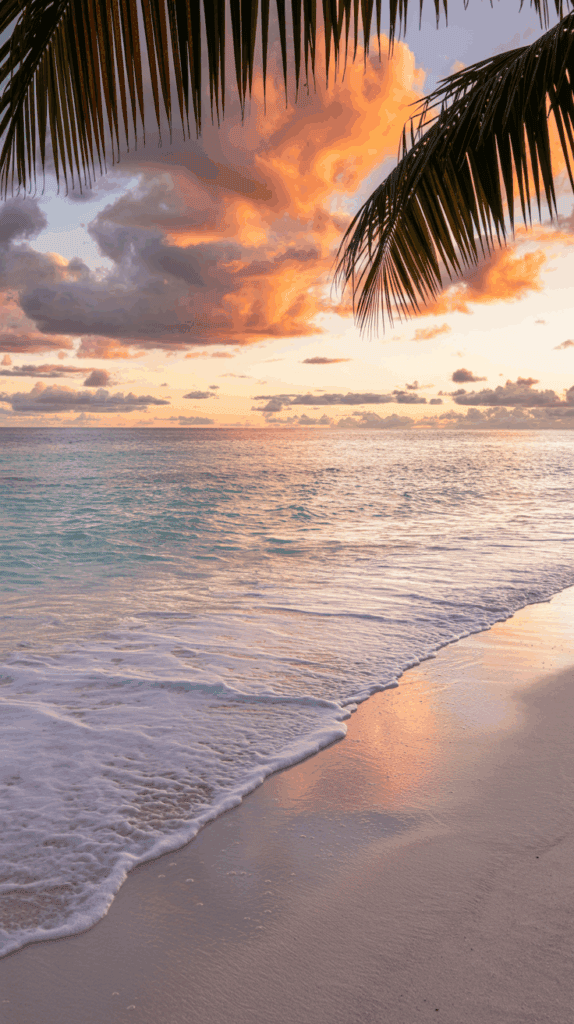 A tranquil beach at sunset with gentle waves lapping the shore, palm fronds framing the scene, and clouds illuminated by the warm glow of the setting sun reflecting on the water.