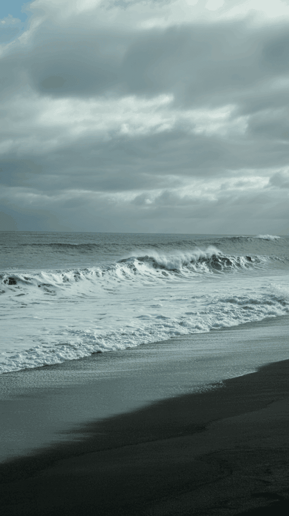 Waves crashing onto a black sand beach under a cloudy sky.